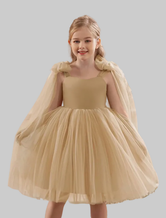 Young girl in a beige dress with a sheer cape standing indoors with floral decorations.