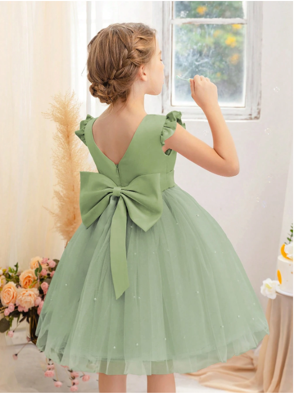 Young girl in a green dress with a large bow standing by a window.